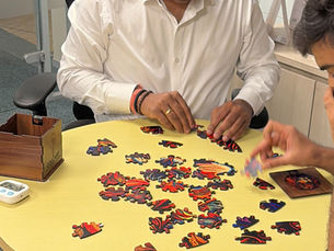 Two people work on a colorful jigsaw puzzle at a round table. One in a white shirt focuses intently. A wooden COASTER box and a timer are nearby.