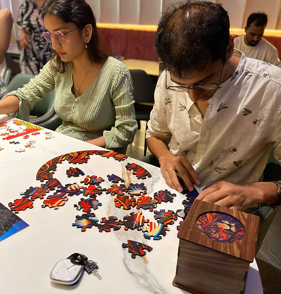 Two adults concentrate on a colorful jigsaw puzzle at a cafe table, with scattered pieces and warm lighting in the background.