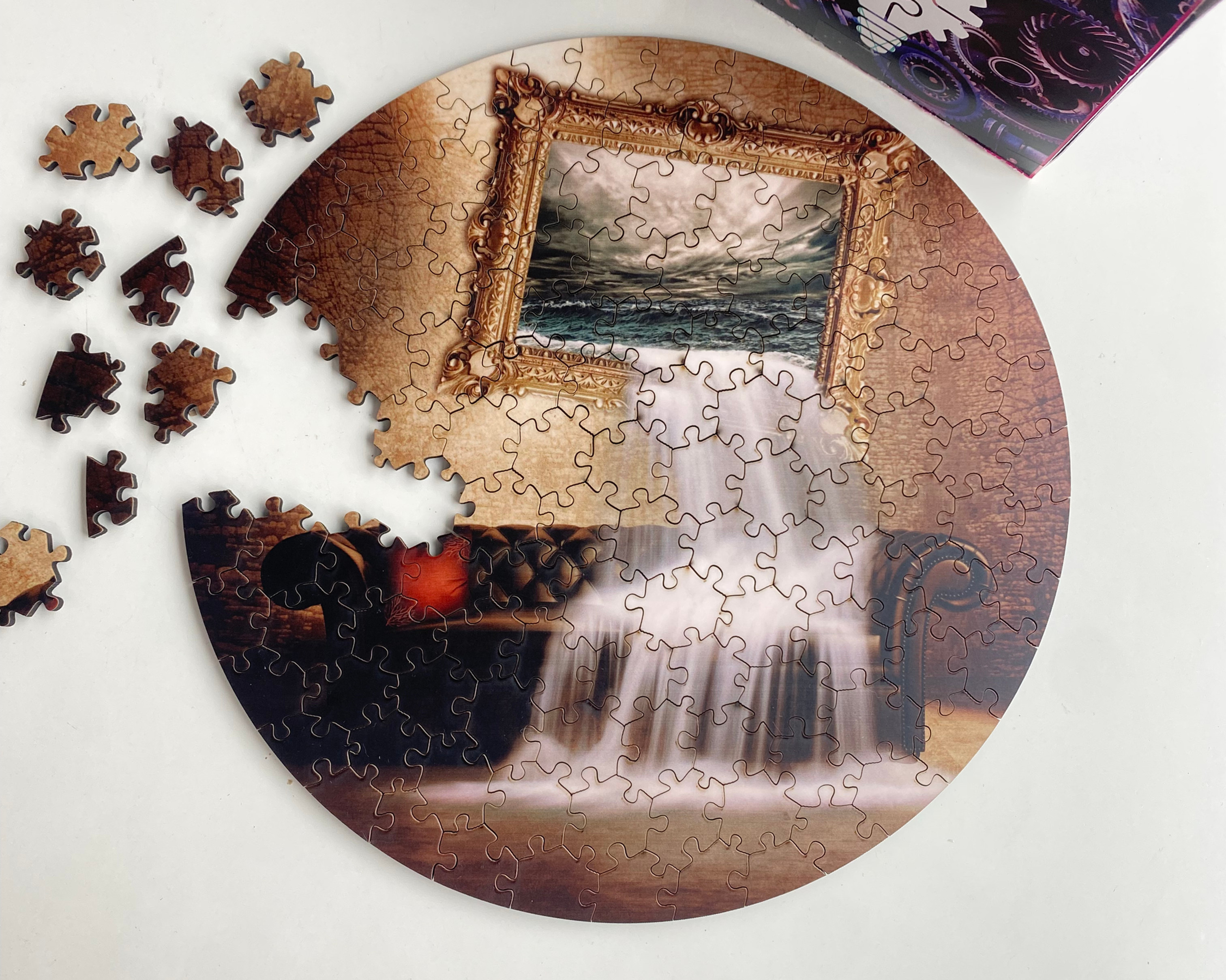 Partially assembled circular jigsaw puzzle on white table, with loose pieces beside it and a colorful gears-pattern box in the corner