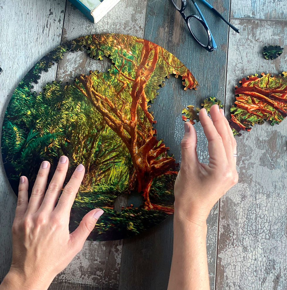 Hands assemble a circular puzzle with a vibrant forest scene on a wooden table. Glasses and a book are nearby.