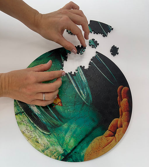 Hands assembling a round jigsaw puzzle with an aquatic image showing a fish and coral. Puzzle pieces are being placed on a white surface.