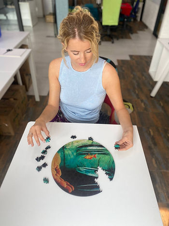 A woman assembles a circular wooden puzzle with an underwater goldfish scene, focused and holding pieces in both hands.