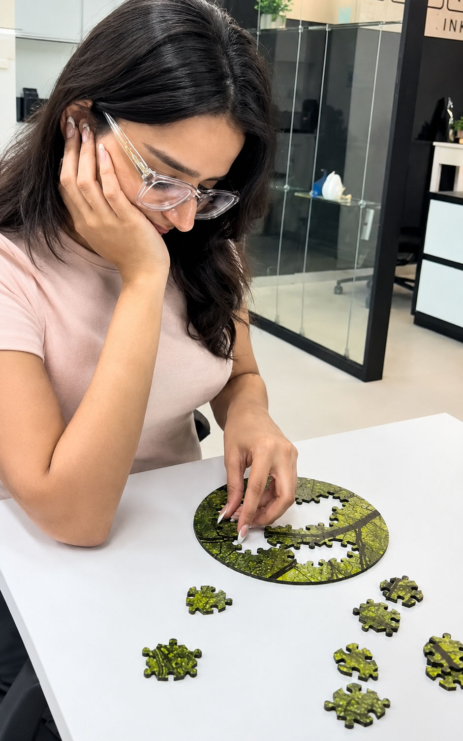 Woman with glasses assembles a green jigsaw puzzle at a white desk in a modern office, looking focused and calm.