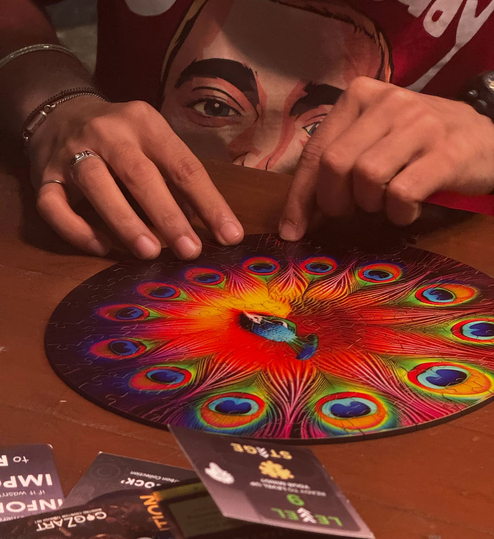 Hands assembling a colorful peacock puzzle on a wooden table. Person wears a red shirt with a face graphic. Cards with text lie nearby.
