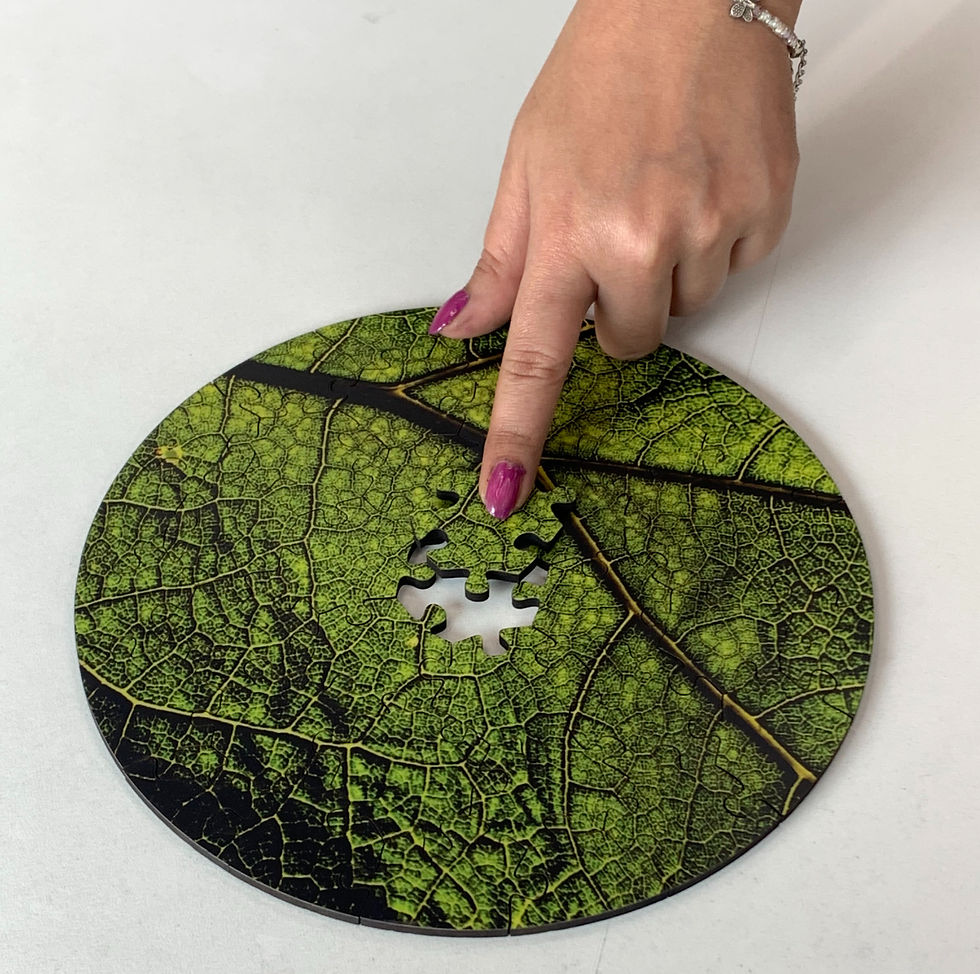 A hand with pink nails completes a green leaf-patterned jigsaw puzzle on a white surface, adding a piece to a round section.