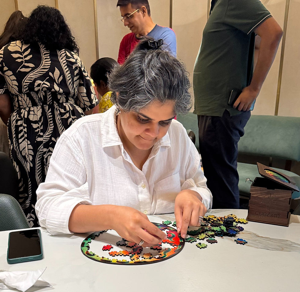 Woman in white shirt assembling a colorful jigsaw puzzle at a table, with others standing behind her and a COGZART box nearby.