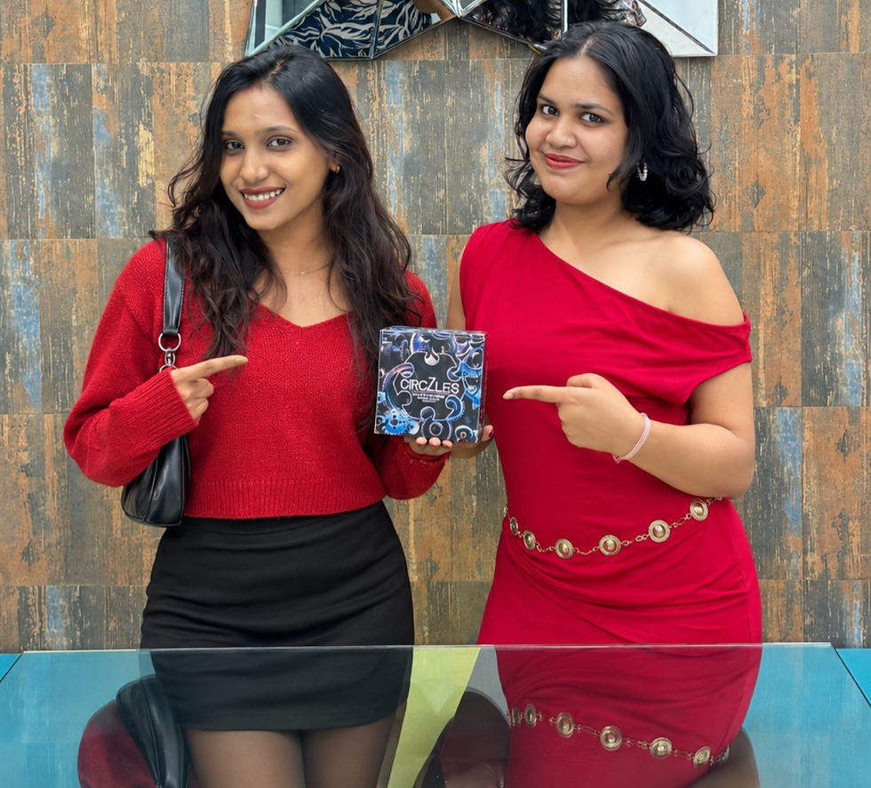 Two smiling women in red outfits point to a CircZLES box, standing before a tiled wall and glass table.