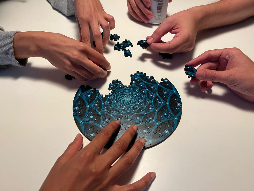 Several hands assemble a blue circular puzzle on a white table, with loose pieces scattered around and a soda can at top.