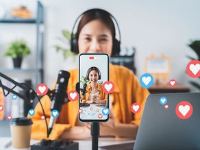 Woman in headphones vlogging at a desk, filmed on a phone tripod with mic, laptop and floating heart icons in a bright home studio.