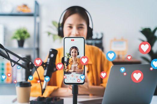 Woman in headphones vlogging at a desk, filmed on a phone tripod with mic, laptop and floating heart icons in a bright home studio.