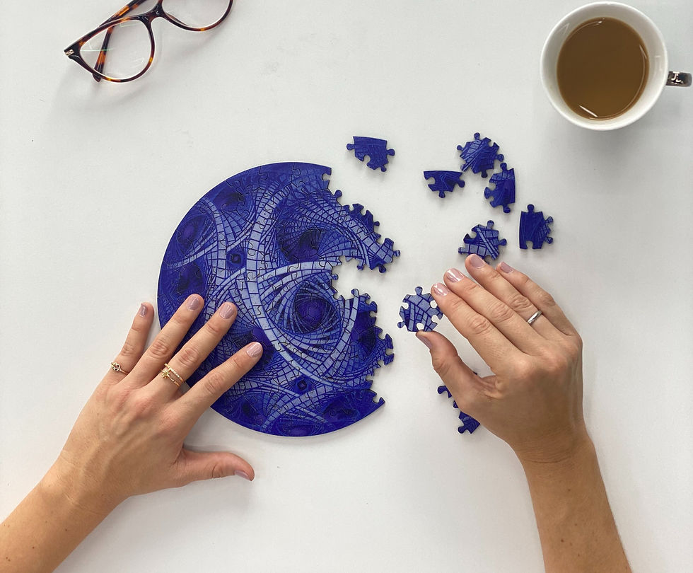Hands assemble a blue patterned circular puzzle on a white table. Nearby, glasses and a cup of coffee suggest a calm, focused atmosphere.