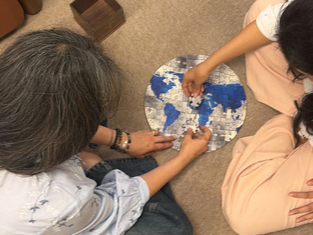 Two people work on a round world map puzzle on a beige carpet. One wears a floral shirt, the other in pink pants. A wooden box is nearby.