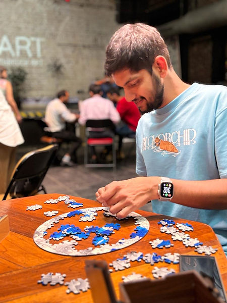 Man in a blue shirt solving a blue and white puzzle on a wooden table. Background shows blurred group sitting. Text "ART" on brick wall.