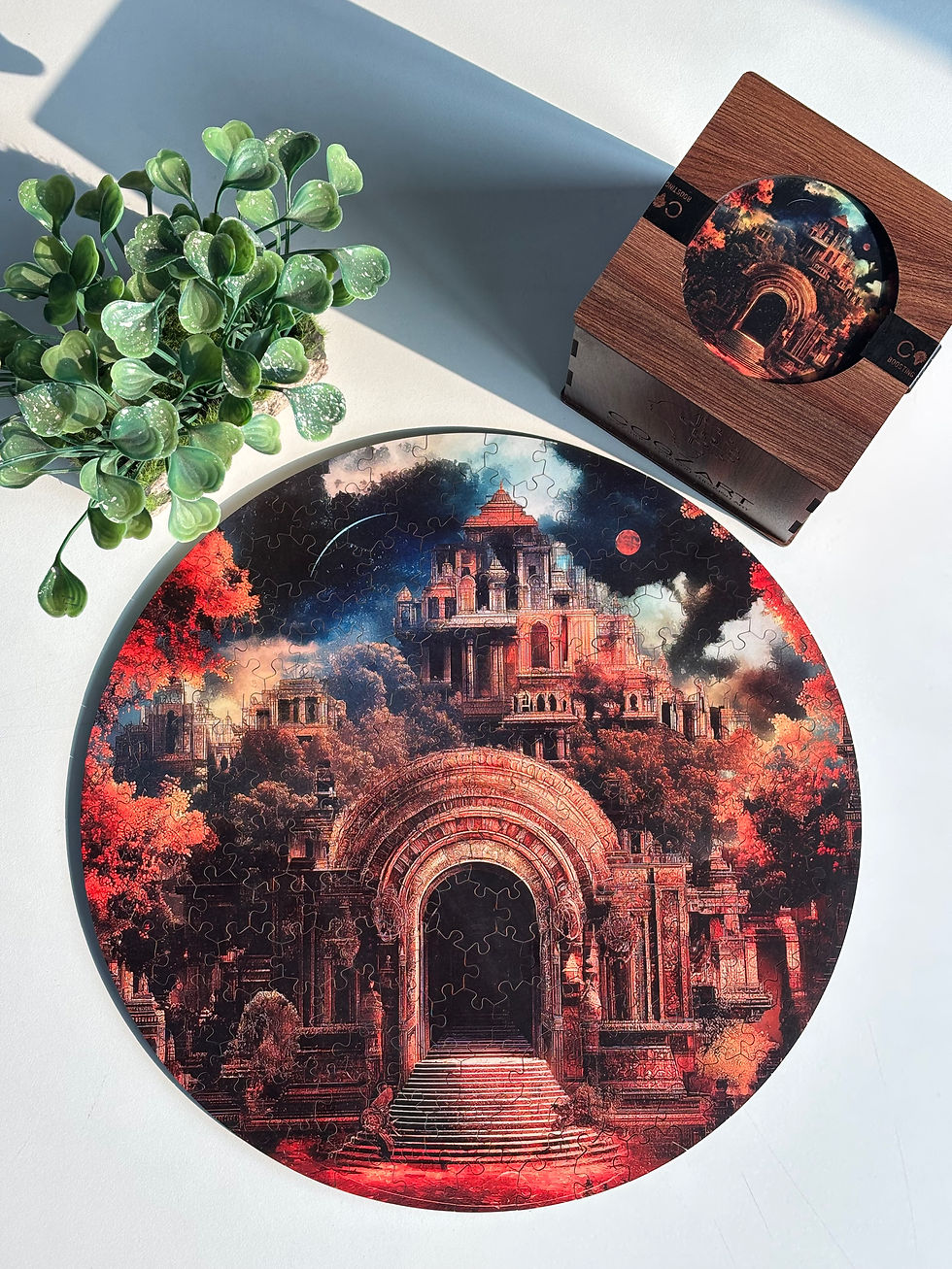 A round puzzle depicting a mystical, ancient temple with red and orange hues sits on a table beside a plant and a wooden box.
