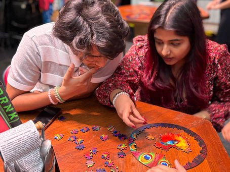 Two people assembling a colorful jigsaw puzzle at a wooden table, focused and engaged. Background is a vibrant, bustling café.