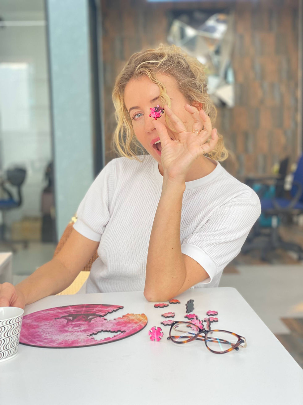 Woman playfully holds a pink puzzle piece over her eye while working on a circular wooden puzzle at a table, with coffee nearby—capturing a cozy, mindful solo moment.