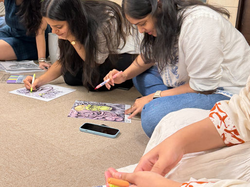 Women coloring detailed affirmative coloring book with markers on a carpeted floor. A green plant in a white pot is visible in the background. Calm mood.