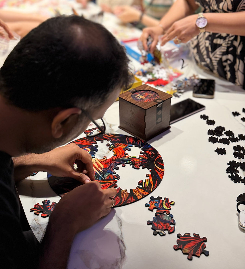 Man assembling a vibrant, circular puzzle on a table with others. A wooden box and scattered pieces are nearby, creating a focused mood.