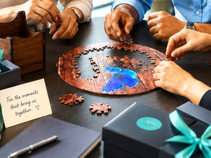 Hands assemble a round jigsaw puzzle at a table, with gift boxes and a note reading For the moments that bring us together. ♥