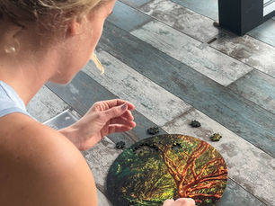 Woman assembling a round puzzle with a tree image on a wooden floor. She's focused, wearing a blue top and floral skirt. Bright setting.
