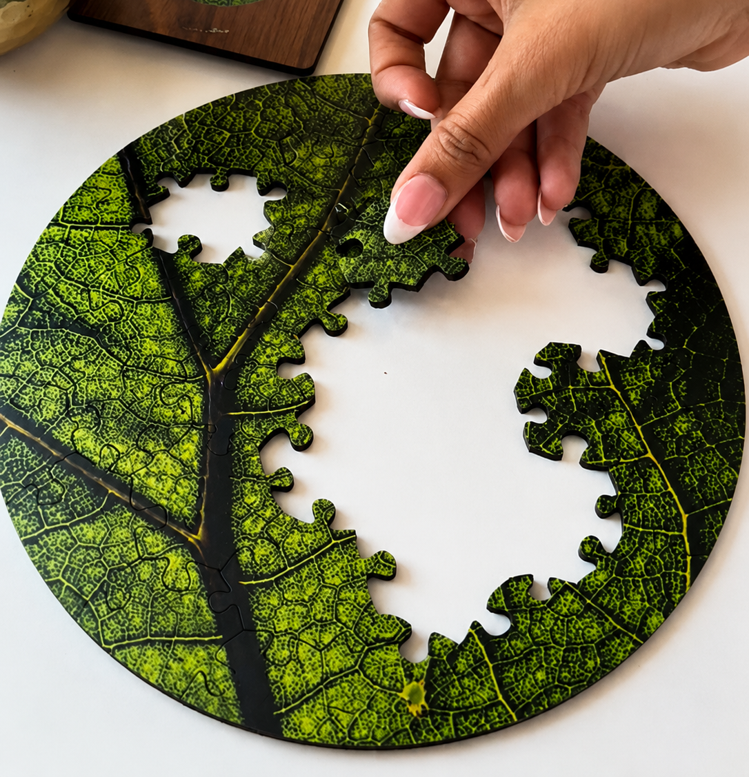Hand placing green leaf-pattern puzzle pieces on a white table beside a wooden tray and potted plant.