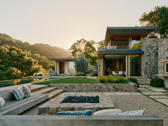 Sunset view across a sunken stone fire pit and gravel terrace toward the stone-clad main house
