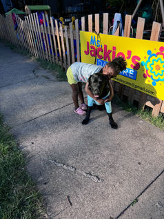 A young child with light brown skin hugs a black dog wearing a light blue shirt while standing on a sidewalk in front of a bright yellow Ms. Jackie’s Daycare sign. A wooden fence and outdoor play area are visible behind them.
