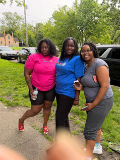 Three women pose together outside on a sidewalk near a grassy yard, parked cars, and trees. Two wear Ms. Jackie’s Daycare shirts, one bright pink and one blue, while the third wears a gray shirt and glasses. One person holds a water bottle, another holds a partially eaten cupcake.