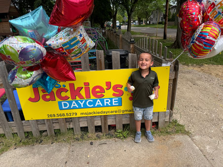 A young child with light brown skin stands smiling beside a bright yellow Ms. Jackie’s Daycare sign decorated with colorful birthday balloons. The child wears a dark green polo shirt, denim shorts, and gray shoes while holding a small treat and a cup. A wooden fence, playground equipment, sidewalk, trees, and a neighborhood street are visible in the background.