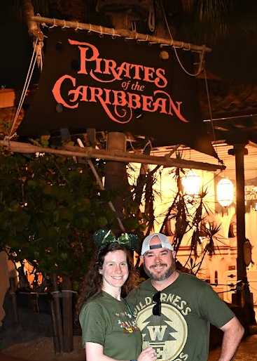 A smiling couple in green shirts stand under a "Pirates of the Caribbean" sign at night, with warm lights and tropical plants in the background.