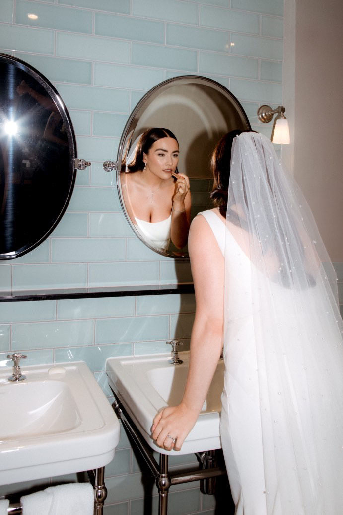A bride in a pearl veil applying lipstick in the mirror in a bathroom

