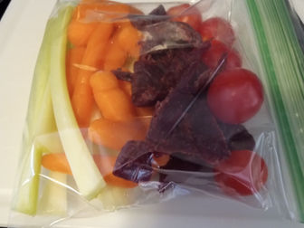 A zip-lock bag with carrots, celery, cherry tomatoes, and beef jerky on a white cutting board. (Vibrant Colors)