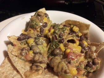 A plate of nachos topped with melted cheese, black beans, corn, and vegetables. The setting is a kitchen with a pan in the background.