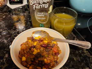 Bowl of chili with corn and beans on a dark marble counter, accompanied by a green juice bottle labeled "Bolthouse Farms Dailey Greens" and a 8 oz. glass of the same juice.