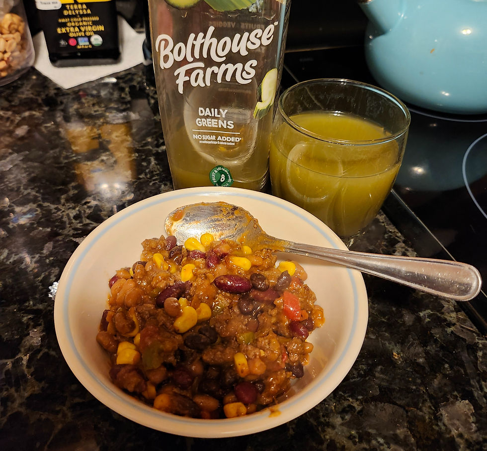 Bowl of chili with corn and beans on a dark marble counter, accompanied by a green juice bottle labeled "Bolthouse Farms Dailey Greens" and a 8 oz. glass of the same juice.