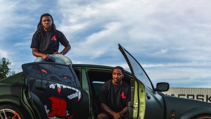 Two people pose with custom green car featuring red logo and monster graphic.