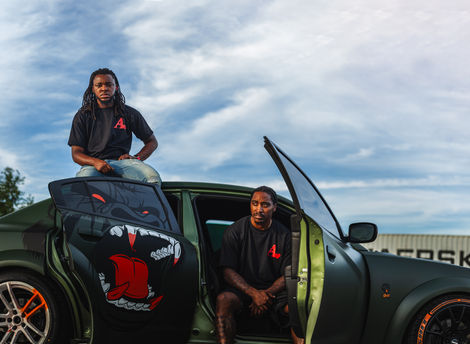 Two people pose with custom green car featuring red logo and monster graphic.