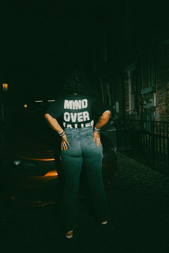 Person in 'MIND OVER MATTER' shirt, jeans, hands on hips, dark street.