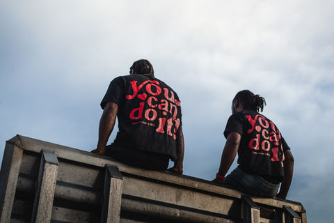 Two men from behind, "You can do it!" shirts, sitting under cloudy sky.