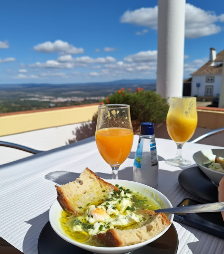 Soup with bread, two orange drinks, and water bottle on outdoor table. Scenic view of landscape under a bright blue sky with fluffy clouds.