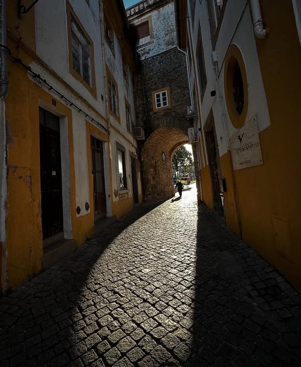 A person walks down a cobblestone alley under an arch, with bright sunlight creating deep shadows. Yellow and white buildings line the narrow path.