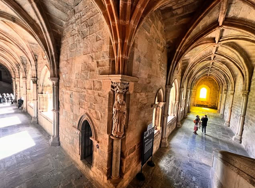 Historic stone hallway with vaulted ceilings. Two people walk towards a glowing arched window. Warm lighting, intricate carvings, serene atmosphere.