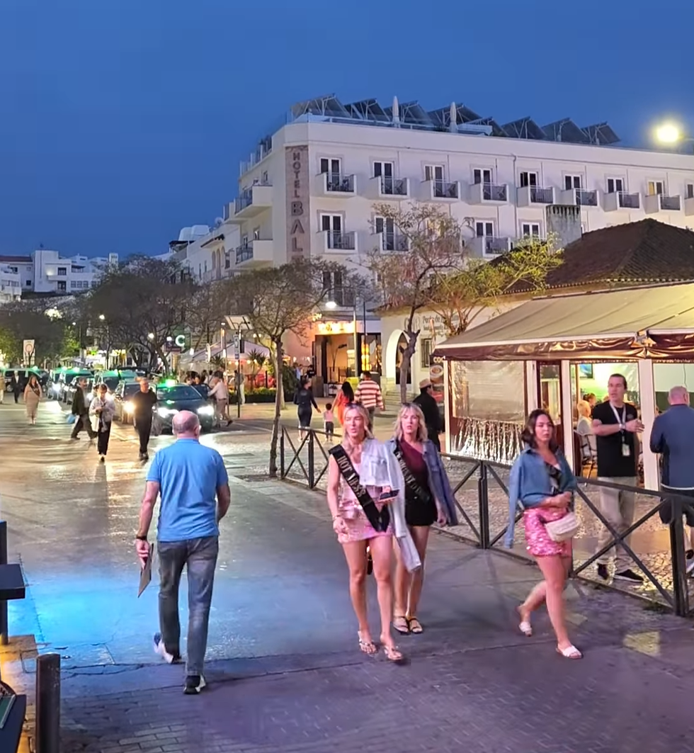 People walk on a bustling, well-lit street at night. A hotel sign reads "Bal" on a white building. Lively and vibrant atmosphere.