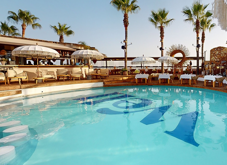 Poolside lounge with umbrellas, tables, and palm trees under a clear sky. Reflections on water; calm, inviting atmosphere.