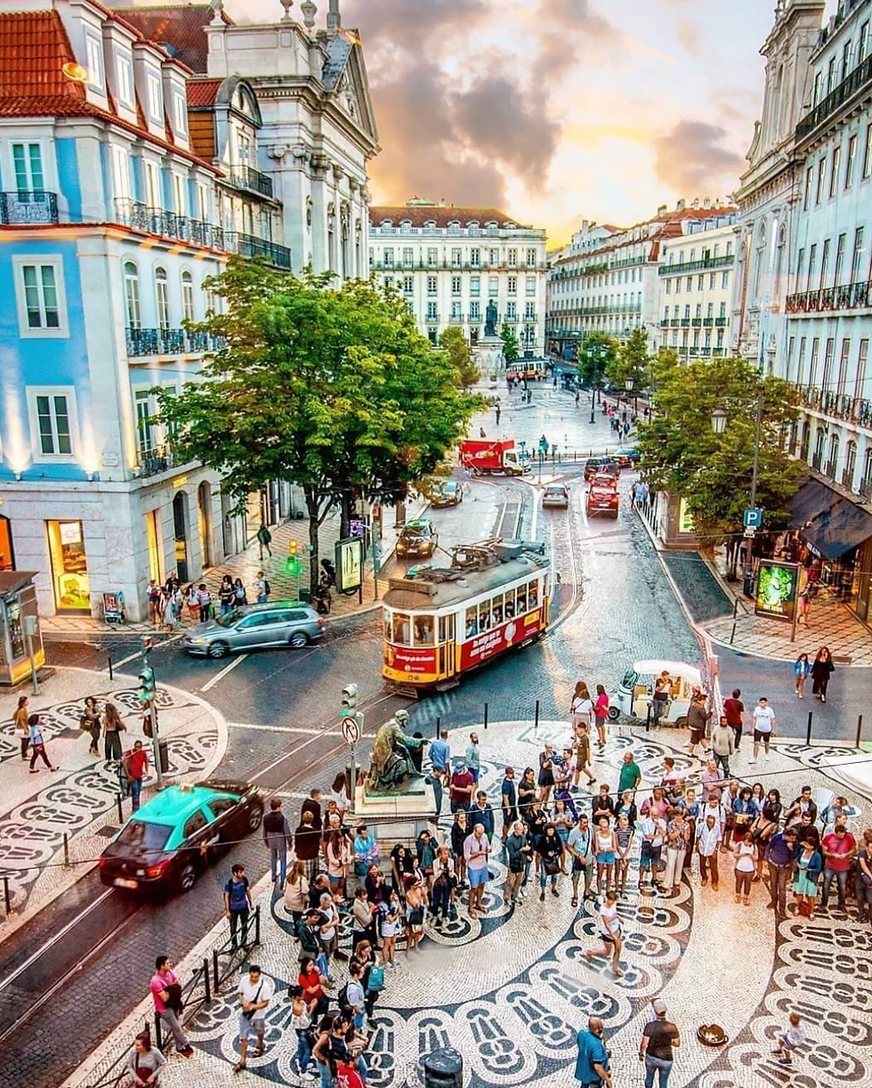 Crowded city square with decorative tiles, people watching a street performer, yellow tram passing, colorful buildings, and sunset sky.