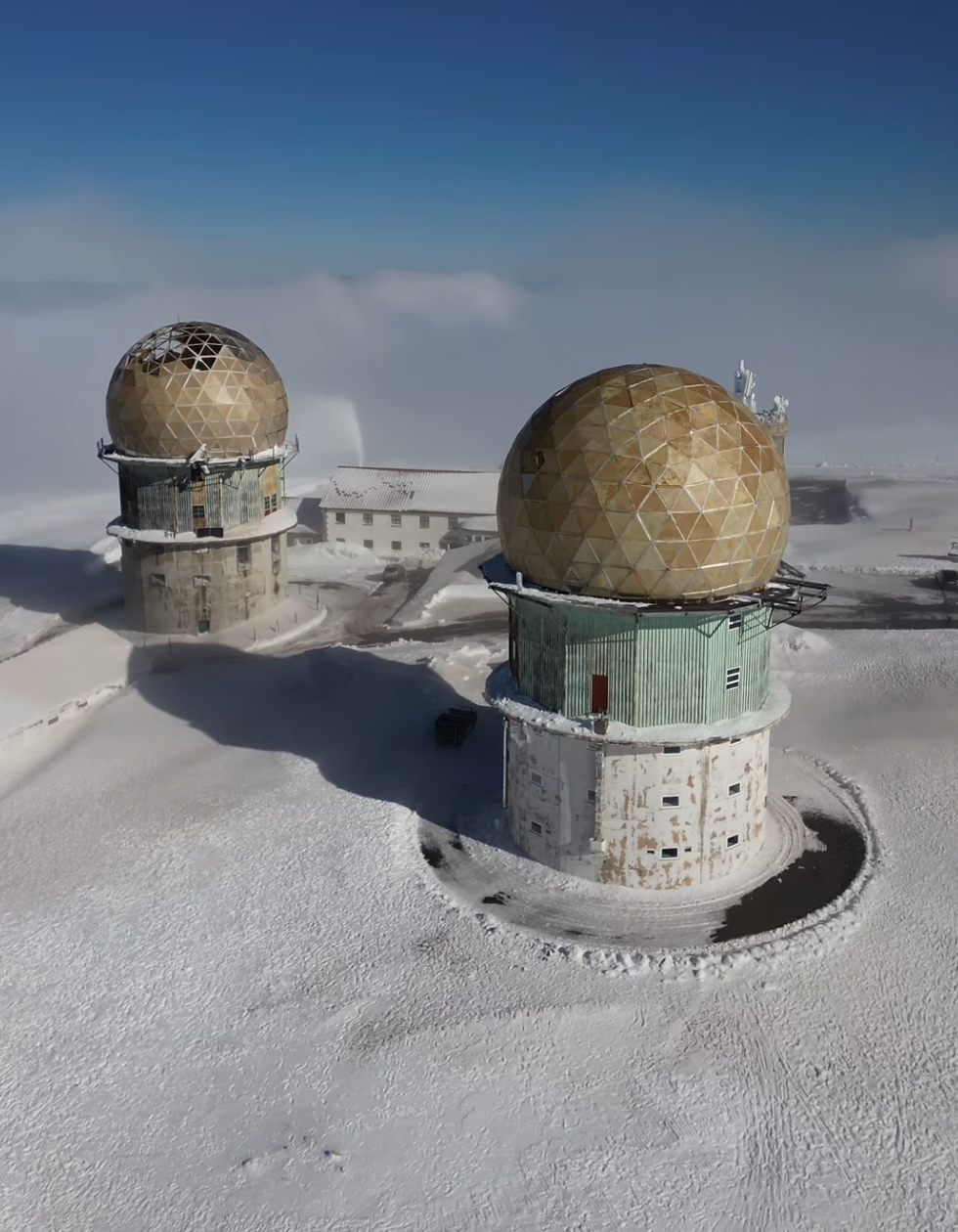 Twin geodesic radomes on snow-covered terrain under clear blue sky. Background includes a building with a white roof.
