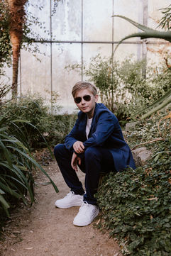 Tienerjongen in een blauw kostuum tijdens communiefotoshoot in de canna banana in Putte, met een zonnebril, zittend op een steen