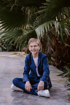 Tienerjongen in een blauw kostuum tijdens communiefotoshoot in de canna banana in Putte, in kleermakerszit op de grond