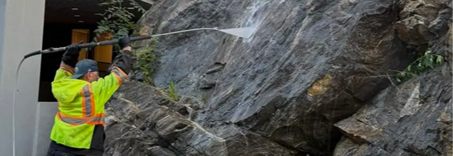 Power washing professional blasting deep mud and dirt buildup off a steep stone wall and rock face.