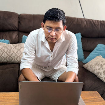 Man in glasses using laptop on a table with a sofa in background.